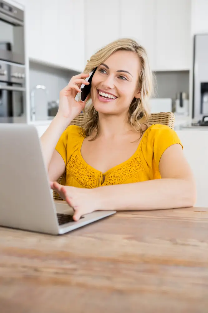 Femme souriante parlant au téléphone tout en utilisant un ordinateur portable, illustrant un service client attentif et disponible.
